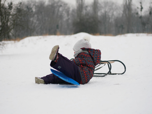 A kid bundled up in a coat, snow pants, boots, and a hat leans against a wooden sled.