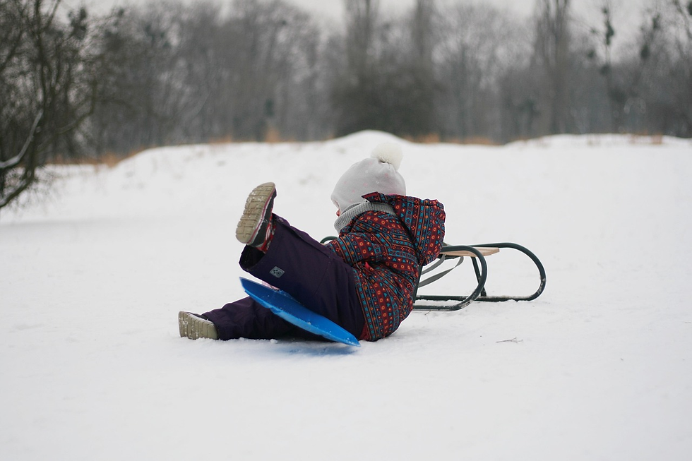 A kid bundled up in a coat, snow pants, boots, and a hat leans against a wooden sled.