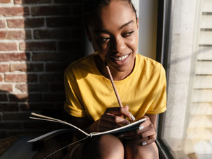 A young woman sits looking out a window with a pencil and notebook in hand
