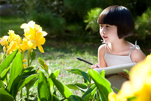 A little girl sits in a garden with a pad of paper and pen