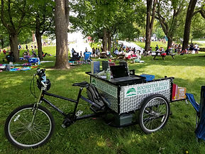 A bike with an attached trailer is parked in a busy park. The trailer has the RHPL logo on its side.