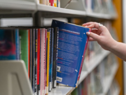 A person reaches in from out of frame to pull a book off a full shelf.