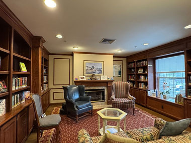 A picture of the mini-branch library at Waltonwood at University, a wood-paneled room with armchairs flanking a fireplace, and a cushioned couch and chair facing it. Books are arranged on built-in shelves on both sides of the room.
