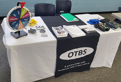 The OTBS table at the last low vision expo. On the right side of the table there are Digital Talking Book Machines and cartridges on display. In the middle is literature about OTBS. On the left side are examples of 3D printed assistive devices, and a prize wheel to spin for swag giveaways. The OTBS logo banner hangs down the middle of the table.