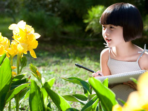 A young girl sits in a garden with a pen and paper, and looks at yellow, fully-bloomed flowers