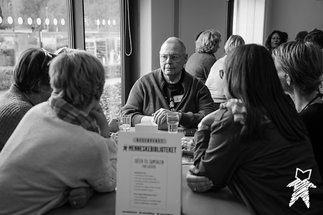 A group of people sit around a table facing a Book (a person leading a discussion about their experiences), who is listening intently to a Reader (a person who is asking questions).
