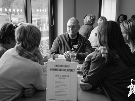 A group of people sit around a table facing a Book (a person leading a discussion about their experiences) who is listening intently to a Reader (a person who is asking questions)