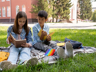 Two kids sit on a blanket outside reading books.
