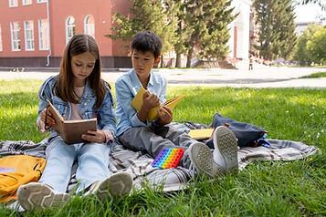Two kids sit on a blanket outside reading books.