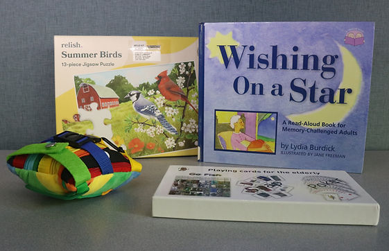 One of the Dementia MYLE Kits available for checkout from Outreach Services. Items shown are a summer bird jigsaw puzzle, a book entitled "Wishing on a Star: A read aloud book for memory-challenged adults," card games, and a buckle pillow - a small square pillow surrounded with various buckles, snaps, zippers, and other fidget items.