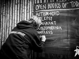 An photo from Human Library. A librarian writes titles of books under the heading "Open Books of Today" on a chalkboard. Book titles include: Autism & ADHD, Epilepsy, Immigrant, Schizophrenia, and Homeless