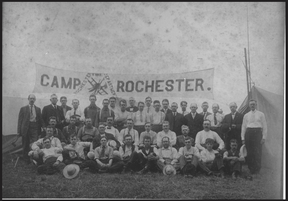 A historical black and white photograph of a group of 40 men arranged in rows for a portrait. The banner behind them reads "Camp Rochester" and has a logo of a helmet, spear, and axe arranged on top of a ladder, surrounded by the words "Rochester Fire Department"