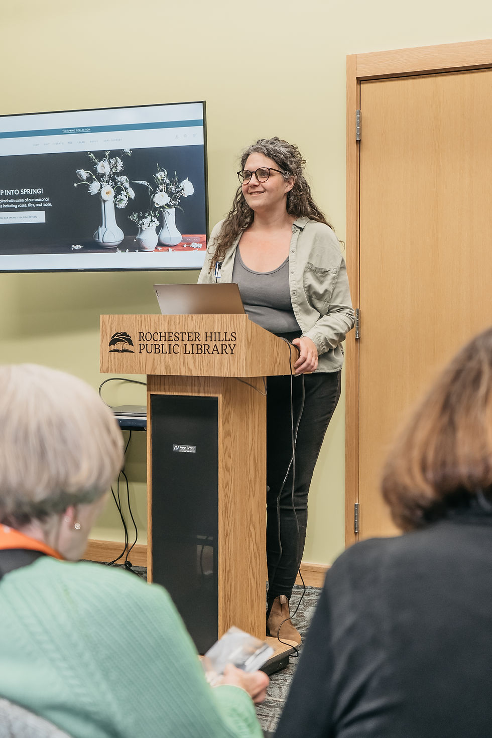 A woman stands behind a podium with Rochester Hills Public Library printed on the front. Over her shoulder is a TV screen displaying the Pewabic Pottery website.