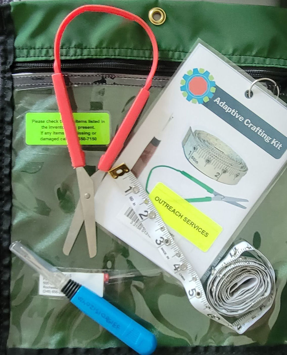 Adaptive scissors, a seam ripper, and tape measure are arranged on top of a card with "Adaptive crafting kit" written at the top, and a circulating bag