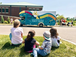 A group of four kids sits in the foreground, looking at the blue bus in the background.