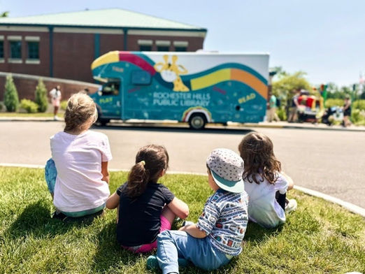A group of four kids sits in the foreground, looking at the blue bus in the background.