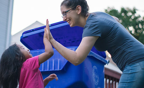 A monther and child high-five in front of a large, wheeled recycling bin.