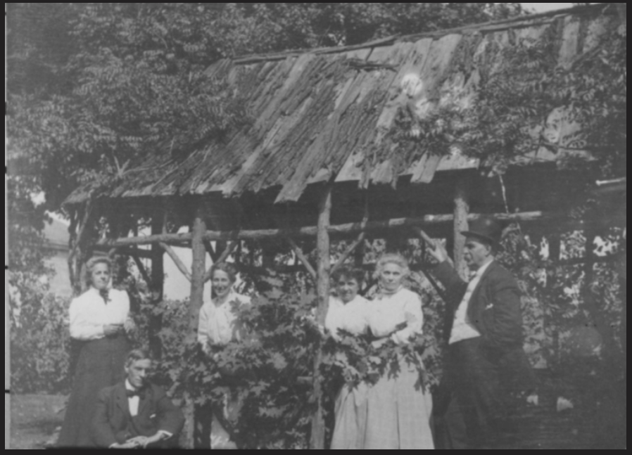Historical black and white photograph of a family of six standing under a covered outdoor wooden structure.