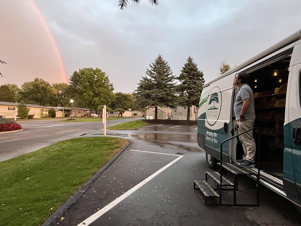 A library employee stands at the open side door of the bookmobile, admiring a rainbow after a rainy day at a bookmobile stop.