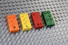 Orange, yellow, red, and green LEGO Braille Bricks on a gray lego platform. The bumps on each brick speall out "PLAY" in Braille