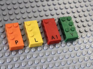 Orange, yellow, red, and green LEGO Braille Bricks on a gray lego platform. The bumps on each brick speall out "PLAY" in Braille