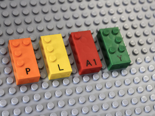 Orange, yellow, red, and green LEGO Braille Bricks on a gray lego platform. The bumps on each brick speall out "PLAY" in Braille