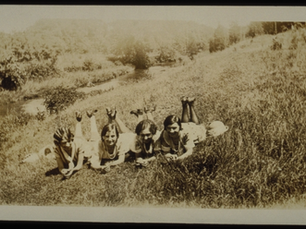 Sepia-toned historical photograph of four women lying on their stomachs on a hill in the grass with their feet up, smiling at the camera.