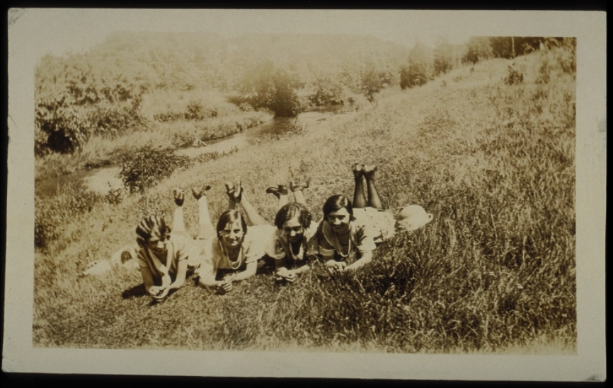 Sepia-toned historical photograph of four women lying on their stomachs on a hill in the grass with their feet up, smiling at the camera.