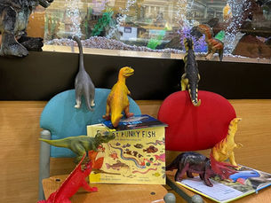 A collection of plastic dinosaurs perched on chairs, books, and the fish tank.
