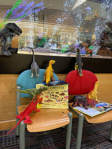 A collection of plastic dinosaurs perched on chairs, books, and the fish tank.
