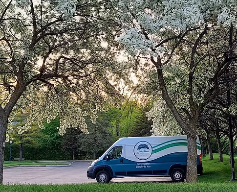 A photo of the RHPL bookmobile parked in the parking lot under an apple tree in full bloom in springtime.