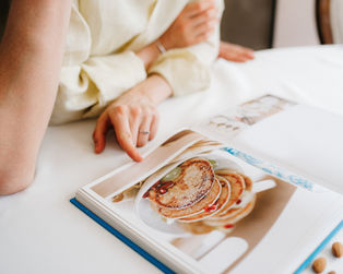 An arm and hand rest on a counter in front of a cookbook that is open to a page with a picture of pancakes.