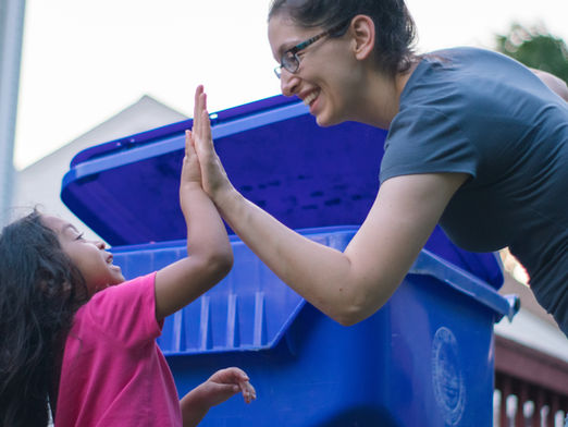 A monther and child high-five in front of a large, wheeled recycling bin.
