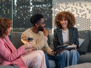 A group of smiling people sit on a couch holding a book, tablet, and coffee.