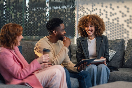 A group of smiling people sit on a couch holding a book, tablet, and coffee.