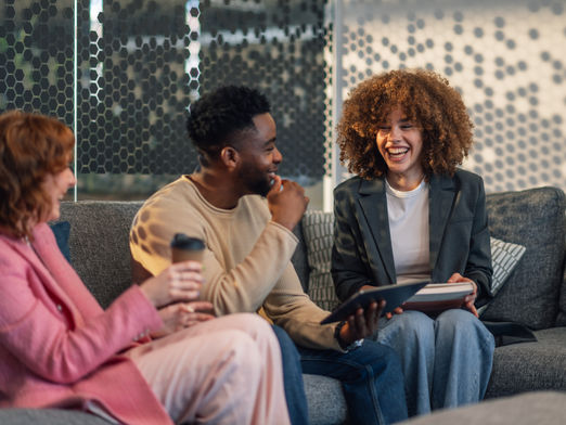 A group of smiling people sit on a couch holding a book, tablet, and coffee.