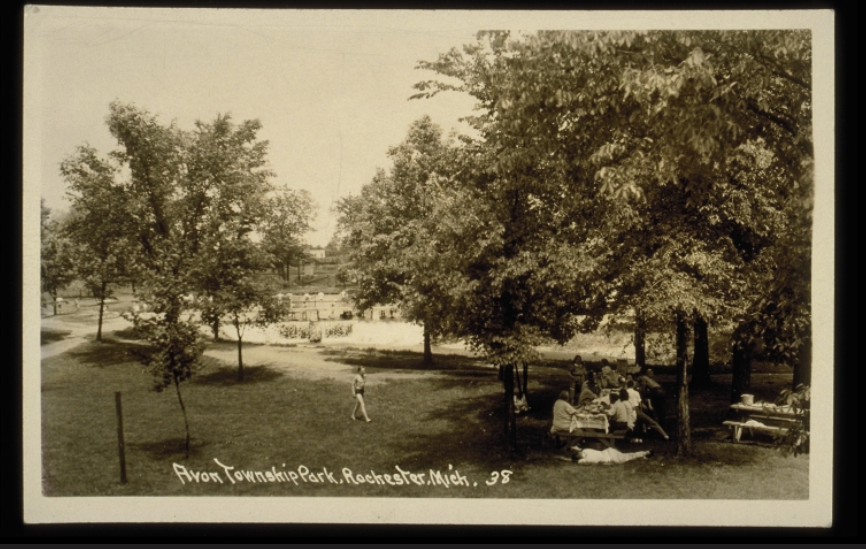 Sepia-toned historical wide-angle photograph of a group of people sitting under a tree. Int he middle of the photo, a man in a bathing suit approaches the group