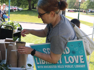 A patron plants a heart leaf philodendron cutting into a biodegradable pot. She holds a sign that says "Spread Library Love" under her arm.