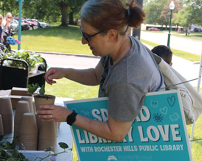 A patron plants a heart leaf philodendron cutting into a biodegradable pot. She holds a sign that says "Spread Library Love" under her arm.