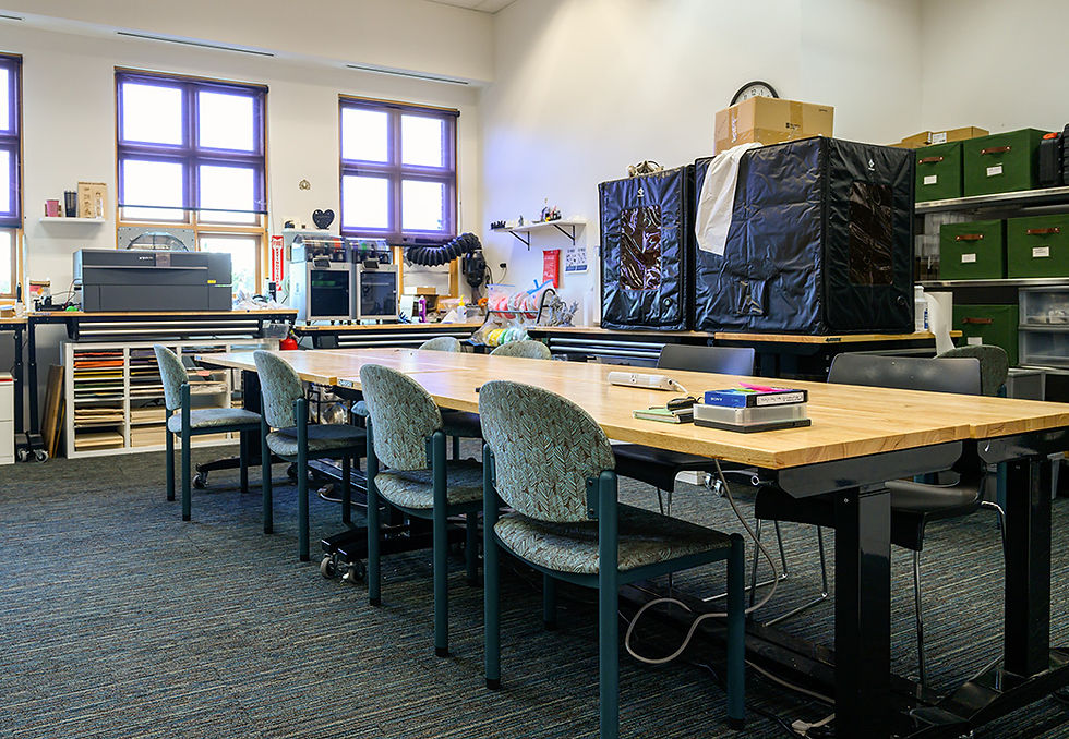 Wooden shop tables sit in the center of the makerspace, in the back there is a laser cutter, two 3D printes, a resin printer, and various supplies for making crafts and projects