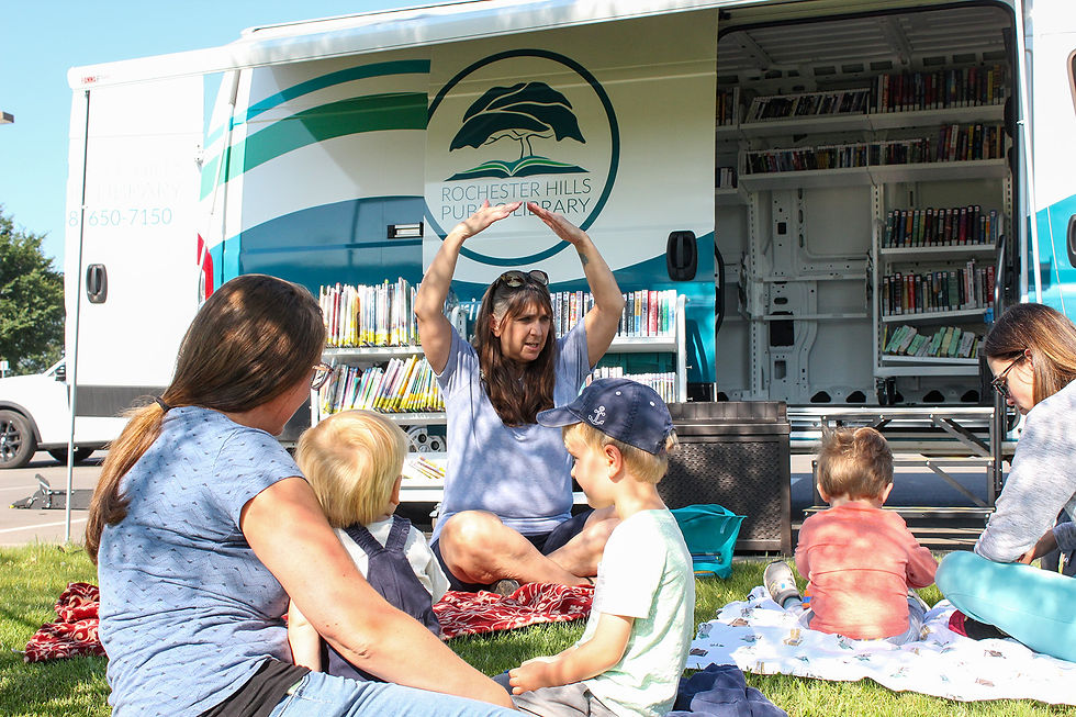 An Outreach Services librarian sits outside of the RHPL bookmobile with her hands over her head as part of an outdoor storytime at the park. A family sits o the grass wachng and singing.