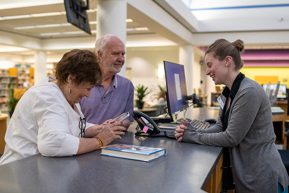 An adult librarian helps two patrons at the Adult Services desk. All three people are smiling at each other.