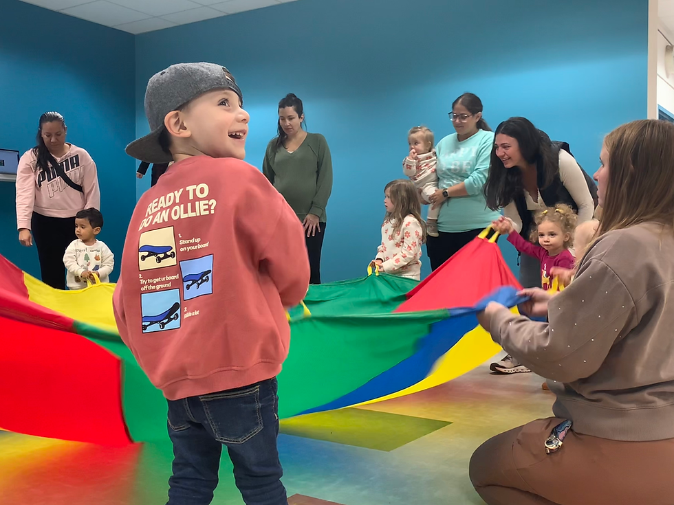 Children and caregivers stand around a large parachute, holding onto the corners. A five year old boy with a backwardss baseball cap smiles at somebody behind him.