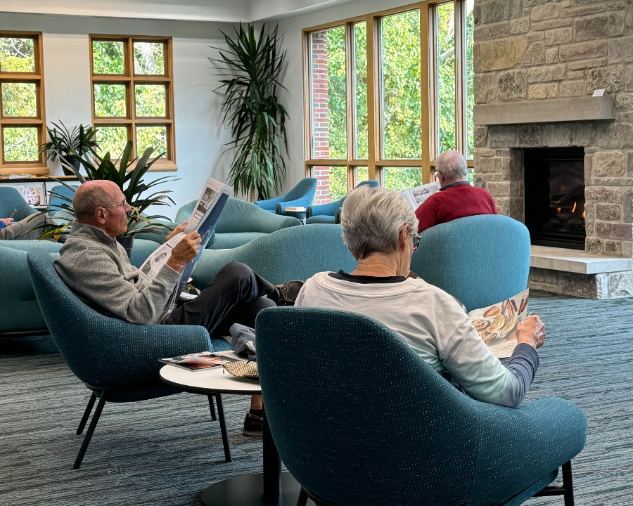 A man and woman sit next to each other by the fireplace reading a newspaper and magazine