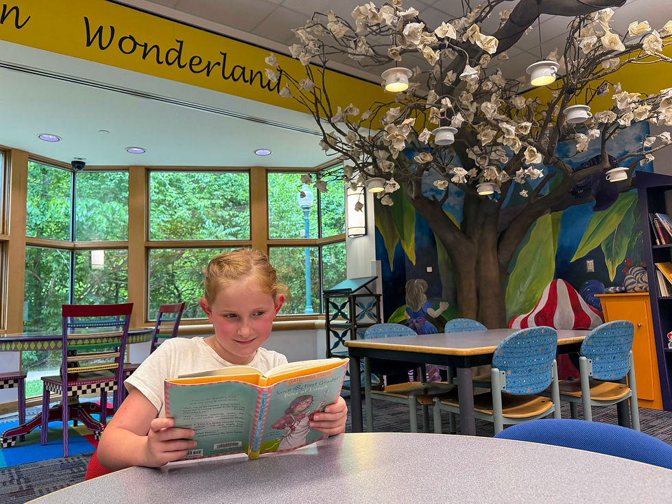 An elementary aged girl reads in the youth room near an Alice in Wonderland themed tree