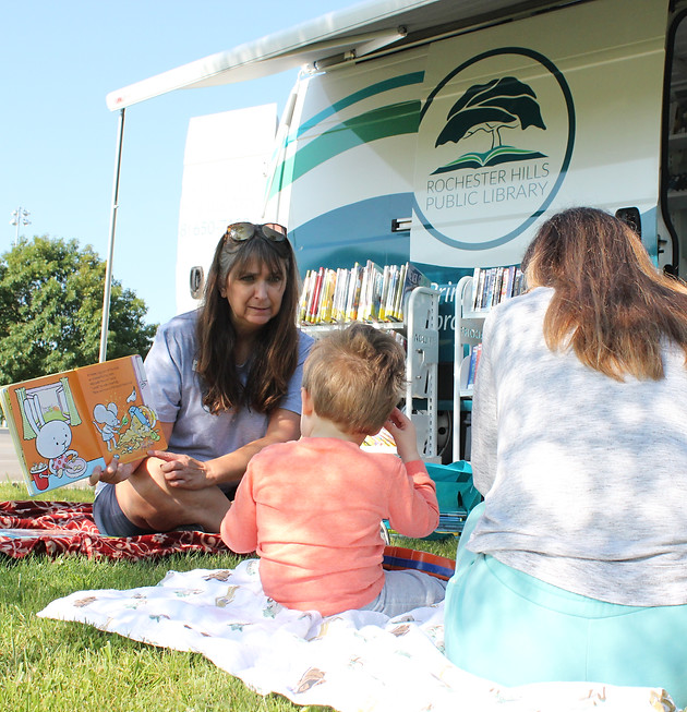 A librarian reads to a child and their caregiver outside by the bookmobile